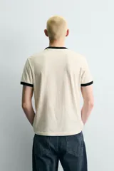 Camiseta de corte regular fit, color crema, con cuello redondo y mangas cortas. Presenta un estampado frontal con un personaje de dibujos animados jugando al baloncesto y texto "Hudson Valley Company Champion '87 NYC, USA". Los acabados del cuello y las mangas son de vivo negro a contraste.