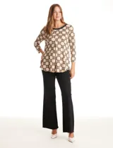 Blusa de seda fría estampada con un patrón geométrico en tonos blanco, negro y marrón. Presenta un escote redondo con detalle de transparencia y un ribete trenzado negro con un aplique dorado. Tiene mangas tres cuartos y un corte fluido con una pequeña tabla central que crea un drapeado suave. El ruedo es redondeado y ligeramente más largo en la parte posterior.