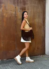 Cartera de hombro tipo hobo confeccionada en cuero gamuzado color chocolate, con detalle de flecos largos en la parte inferior y correa ajustable con hebilla metálica.