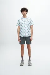 Camisa de manga corta con estampado de flores y palmeras en tonos azules, verdes y naranjas sobre fondo blanco. Tiene cuello americano, bolsillo en el pecho y botones personalizados.