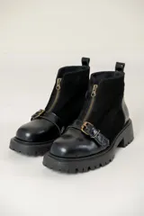 Bota corta de cuero negro con suela track, cierre frontal con cremallera y tira decorativa con hebilla metálica sobre el empeine.