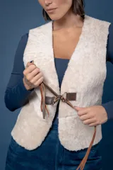 Chaleco de piel sintética en color beige, con cierre frontal mediante hebillas metálicas y tiras de cuero marrón que cuelgan como detalle decorativo.