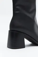 Bota corta de caña elástica, color negro con acabado charolado. Presenta puntera cuadrada y taco medio geométrico y ancho.