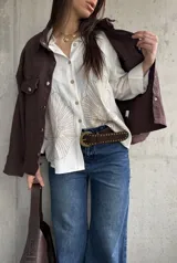 Camisa oversize blanca con botones frontales, cuello camisero y manga larga. Presenta grandes bordados en hilo beige con diseño de hojas o abanicos en la parte inferior frontal.