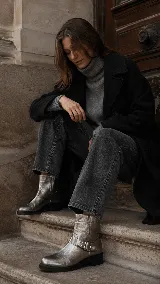 Bota corta de cuero color peltre metalizado, con diseño de caña media, cierre lateral y detalle de correa con hebilla y tachas metálicas en el tobillo. Cuenta con suela plana y taco bajo.
