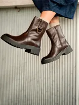 Bota de caña media color chocolate, confeccionada en cuero con suela track de goma negra. Presenta un detalle de argolla metálica en el lateral externo y cierre funcional en el lateral interno.