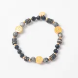 Pulsera elástica compuesta por cuentas de piedra natural gris, cuentas de cristal facetado negro, discos de madera y dijes circulares dorados con diseño de árbol de la vida grabado.