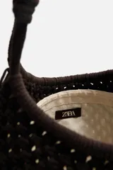 Bolso tipo shopper de tejido macramé marrón oscuro, con forma trapezoidal y asas de hombro trenzadas.