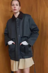 Chaqueta encerada de corte recto con cuello de solapa en pana marrón. Presenta manga larga, bolsillos delanteros de vivo y de plastrón con solapa, y un cinturón de lazada en el mismo tejido. Incluye cierre frontal con botones ocultos por tapeta y abertura en la espalda.