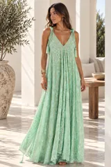 Vestido largo color dorado, confeccionado en tela semitransparente con diseño bordado floral y detalles de brillo. Presenta escote en V, tirantes finos ajustables con borlas en los hombros, y corte amplio y fluido con gran movimiento.
