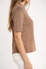 Sweater de tejido de punto fino en color arena, con cuello redondo y mangas cortas. Presenta un diseño minimalista con un pequeño detalle metálico en el ruedo.