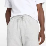 Short deportivo gris melange de algodón y poliéster reciclado, con cintura ajustable con cordón, bolsillos laterales y bolsillo trasero. Logo de New Balance estampado en blanco en la pierna izquierda.