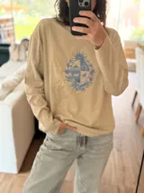 Sweater de punto color beige con cuello redondo y mangas largas. Presenta un escudo bordado en tonos azules en el centro del pecho.