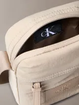 Bandolera Calvin Klein color beige confeccionada en nylon reciclado. Presenta correa ajustable, cierre de cremallera, asa superior y parche con el monograma CK en el frente.
