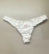 Bombacha tipo less de algodón blanco con bordes fruncidos y un pequeño corazón bordado en hilo rosado en el centro.