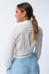 Camisa blanca de corte entallado con cuello clásico, cierre frontal de botones y mangas largas con puños elásticos. Presenta un diseño ajustado en la cintura con una banda elástica integrada que realza la silueta.