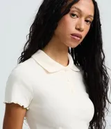 Blusa polo corta de algodón con elastano, color marfil, de manga corta con borde tipo frufrú, cuello camisero y cierre frontal con tres botones.