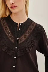 Camisa de manga larga color negro, con cuello redondo y cierre frontal de botones. Presenta un diseño con volados en la parte superior delantera y detalles de encaje bordado en los hombros y pecho.