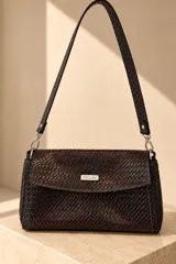Cartera tipo bandolera de cuero vacuno color marrón oscuro con textura trenzada, solapa con broche magnético y placa metálica con el logo de la marca en el centro. Cuenta con correa de hombro ajustable y desmontable unida mediante argollas y mosquetones metálicos.
