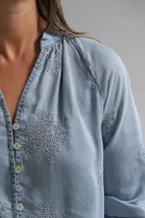 Blusa de tela tipo denim liviano en tono azul claro, con cuello mao, cierre frontal de botones y mangas largas con puños. Presenta un delicado bordado floral en todo el frente y las mangas.