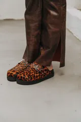 Mules de cuero con estampa animal print de leopardo, suela plana negra y tira ajustable con hebilla metálica y ojales.