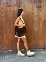 Cartera de hombro tipo hobo confeccionada en cuero gamuzado color chocolate, con detalle de flecos largos en la parte inferior y correa ajustable con hebilla metálica.