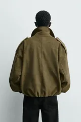 Cazadora cropped fit color verde militar, confeccionada en tejido efecto ante. Cuenta con cuello solapa, manga larga con puño abotonado, trabillas en los hombros, bolsillos laterales ocultos y un bolsillo interior. El bajo tiene elástico interior y el cierre frontal es con cremallera oculta por solapa con botones a presión.