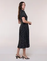 Vestido midi negro con estampado de lunares blancos, de la marca Sharagano. Presenta cuello con solapa, escote en V, cierre frontal con botones y mangas cortas. Tiene un corte en la cintura con lazo ajustable y falda plisada.