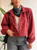 Chaqueta corta de efecto gamuza en color burdeos, con cierre frontal y botones. Presenta mangas tipo globo con puños elásticos y una solapa decorativa con aros metálicos. Incluye ajuste lateral en la cintura y está completamente forrada.