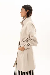 Trench corto color beige con cuello alto, cierre frontal de botones y cinturón ajustable en la cintura. Presenta mangas largas con detalles de tiras ajustables en los puños y bolsillos laterales.