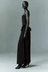 Vestido largo de color negro con escote recto y tirantes finos ajustables. Presenta un diseño con detalle de volados en la cintura que genera un efecto peplum y una falda con caída fluida.