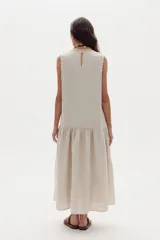 Vestido midi de lino color beige, sin mangas, con cuello redondo y detalles de ondas en las sisas. Presenta un corte con volante fruncido en la parte inferior.