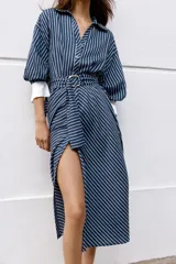 Vestido midi azul marino con rayas blancas, cuello solapa, escote en V y cierre frontal con botones. Cuenta con mangas abullonadas por debajo del codo con puños anchos a contraste, cinturón ajustable del mismo tejido con hebilla metálica y falda evasé.