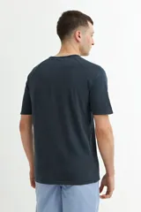 Remera de manga corta color azul oscuro o negro deslavado, con cuello redondo y corte raglán. Presenta un pequeño estampado en relieve o tono sobre tono en el pecho con texto y un símbolo.