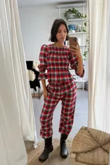 Conjunto de dos piezas confeccionado en viyela con estampado de cuadros escoceses en tonos rojo, negro y blanco. La parte superior es una blusa de manga tres cuartos con frunces en el pecho y puños con volados. El pantalón es de corte holgado con cintura elástica y cordón ajustable.