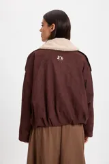 Campera bomber de gamuza color chocolate con cuello de corderito sintético desmontable en color crema. Presenta un diseño holgado con cierre frontal, hombreras decorativas y bordado sutil en el pecho.