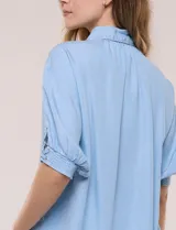 Camisa de tencel liviano estilo denim, color celeste, con cuello camisero, cierre frontal con botones nacarados, mangas cortas abullonadas con puños elásticos en punto smock y detalles de ojalillos.