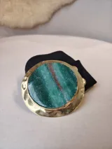 Cinturón de cuero artesanal color marrón con hebilla redonda de metal dorado y piedra natural verde con una veta rojiza. El cinto viene con una funda de tela negra.
