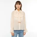Camisa vaporosa color beige claro, con cuello alto y lazo al cuello, mangas largas abullonadas con puños elásticos.