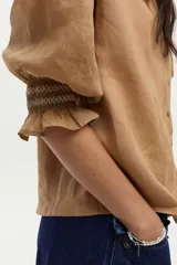 Blusa color tostado confeccionada en lino, con mangas abullonadas de ramie y detalles de bordado en punto smock en los puños.