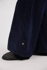 Pantalón sastrero de tiro medio confeccionado en pana de algodón. Presenta un diseño de pierna ancha con costuras verticales decorativas, bolsillos delanteros y traseros, y un detalle de aplique con ojalillo metálico en el ruedo de una de las piernas.