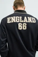 Polo de manga larga en tejido técnico acanalado color negro, con cuello solapa y abertura frontal. Presenta puños con ribete a contraste y una etiqueta aplicada en el pecho con el diseño conmemorativo de la Copa Mundial de la FIFA 1966.