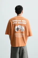 Camiseta boxy fit de punto tejido en hilatura de algodón, color terracota, con cuello redondo y manga corta. Estampado frontal de dados en tonos grises y blancos dentro de un óvalo.