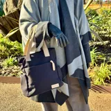 Cartera tipo tote de lona color marrón con asas de mano en color beige. Cuenta con un bolsillo frontal, bolsillos laterales y un detalle de tirador de cierre en cordón blanco.