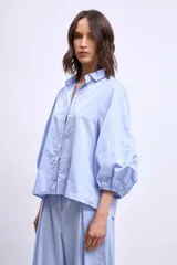 Camisa de algodón color celeste con mangas abullonadas y detalles de bordado lineal blanco en las mangas. Presenta un corte holgado y cierre frontal con botones.