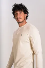 Remera de manga larga tipo Henley, color beige, con cuello redondo y tapeta con botones. Presenta un pequeño logo bordado en el pecho.