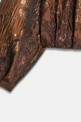 Chaqueta estilo bomber de cuello redondo y manga larga, confeccionada en tejido jacquard con acabado metalizado brillante. Presenta diseño con pliegues y cierre frontal con cremallera.