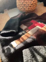 Buzo de lana uruguaya con diseño patchwork geométrico inspirado en el Tetris. Presenta una combinación de texturas y colores en tonos marrón, negro, rojo y gris, con una etiqueta de cuero en la parte inferior trasera.