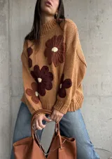 Sweater de punto grueso color camel, con diseño de flores grandes aplicadas en tonos marrones y centro crema. Presenta cuello redondo alto, mangas largas con hombros caídos y terminaciones acanaladas.