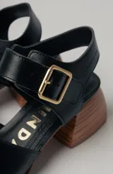 Sandalias de cuero negro con diseño minimalista y tacón ancho geométrico en tono madera. La capellada lisa aporta una estética depurada y moderna, mientras que la pulsera con hebilla metálica ajustable asegura un calce firme y confortable.